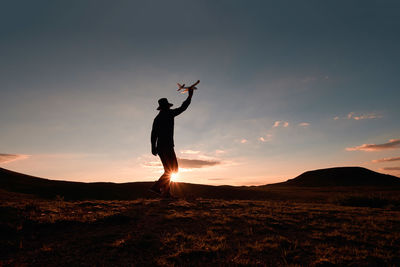 Silhouette man holding model airplane while walking on landscape against sky