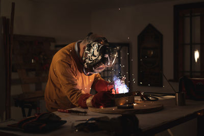 Unrecognizable male employee in protective gloves and helmet using welding machine while working in dark workshop