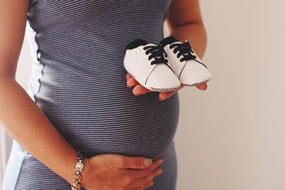 Midsection on pregnant woman holding shoes while standing against wall