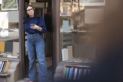 Thoughtful woman holding phone while standing at entrance of bookstore