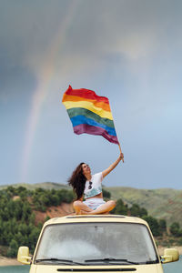 Smiling woman holding rainbow flag sitting cross-legged on van