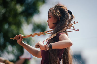 Side view of young woman with sticks standing against sky