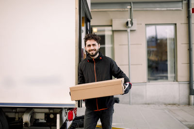 Portrait of confident young delivery man with box leaning on truck