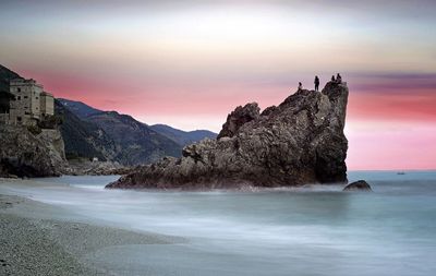 Distant view of friends on cliff at mediterranean sea against sky during sunset