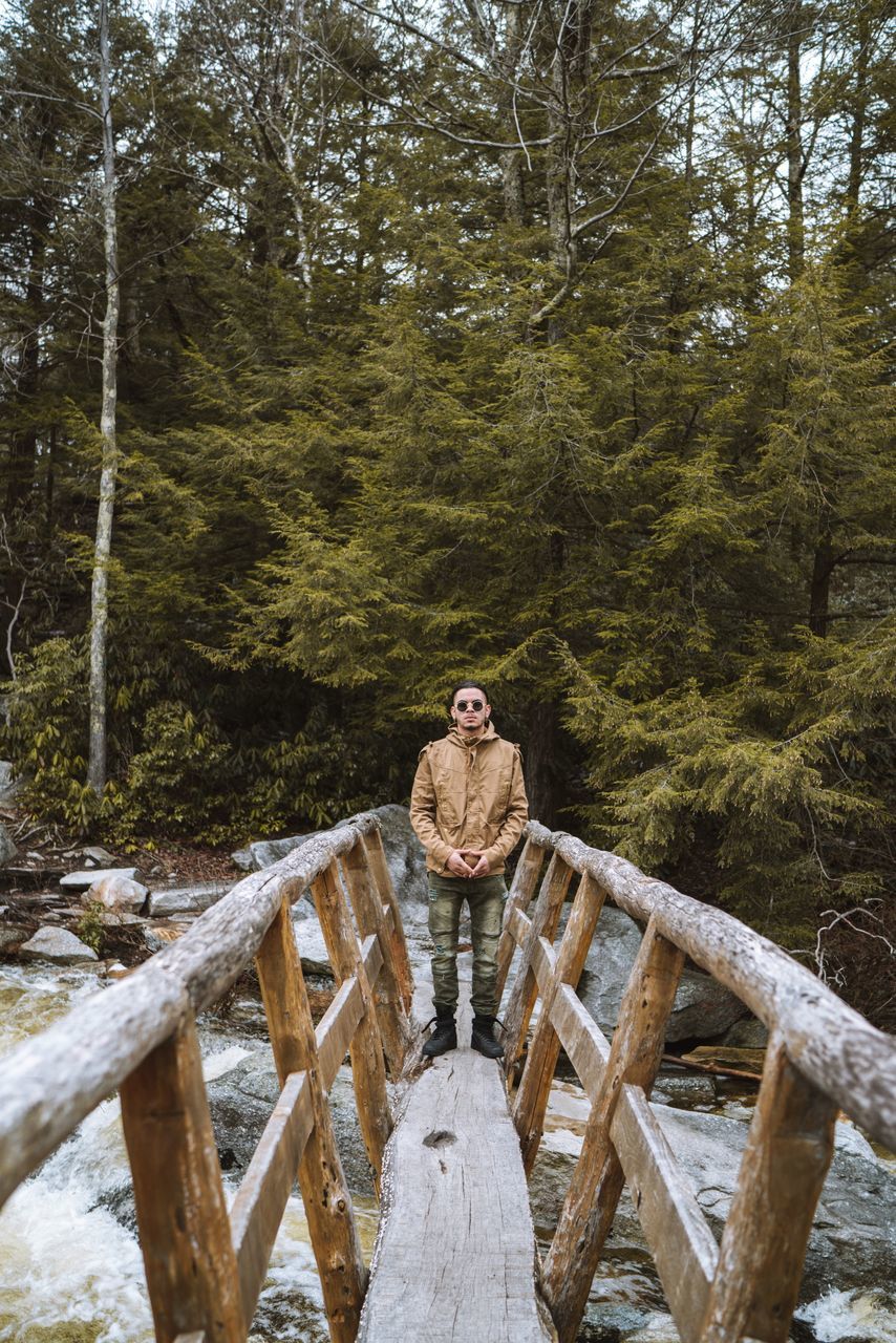 tree, forest, plant, one person, land, real people, leisure activity, nature, young adult, front view, wood - material, day, log, standing, water, full length, adult, beauty in nature, wood, footbridge, woodland, outdoors