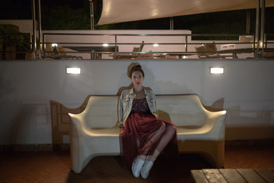 Portrait of woman sitting on bench by illuminated restaurant