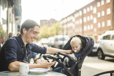 Smiling man listening music through headphones from smart phone while sitting with baby at sidewalk cafe in city