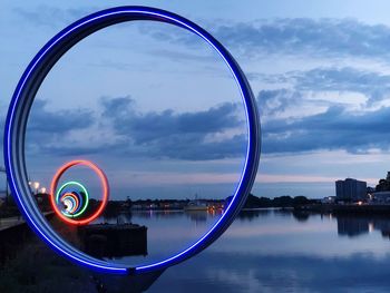 Illuminated rings by river in city against sky during sunset