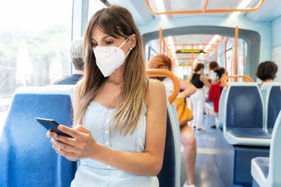 Woman wearing mask using smart phone sitting in bus