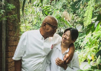 Senior couple with arm around standing against plants