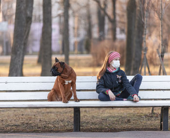 A masked girl and a muzzled dog sit on a park bench and look in different directions