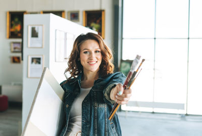 Charming young woman artist in denim jacket with paint brushes and painting on canvas 