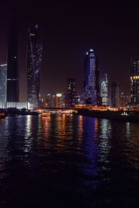 Illuminated buildings by river against sky at night