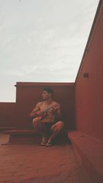 Young shirtless man playing ukulele on building terrace against sky