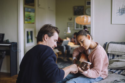 Father teaching daughter to play guitar while sitting on bed at home