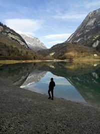 Rear view of person standing at lakeshore against mountains