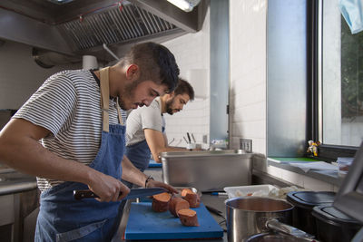 Side view of young hispanic male cook in casual clothes and apron cutting sweet potato while working in restaurant kitchen near colleague