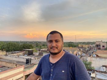 Portrait of young man standing against cityscape during sunset