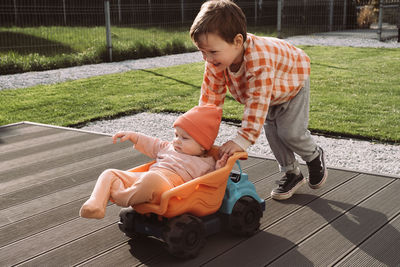 Brother giving a ride to baby sister in big toy car outdoors. children having fun near house. boy