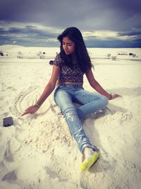 Teenage girl sitting at sandy beach against sky