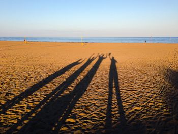 Long shadows of friends at sandy beach against sky
