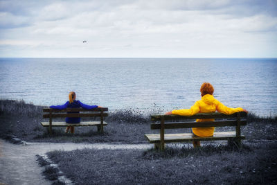 Rear view of people sitting on bench by sea against sky