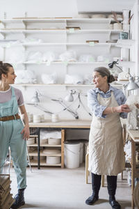 Senior female potter working with young employee against shelves at workshop