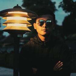 Young man standing by illuminated lamp post at dusk