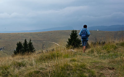 Rear view of young man with backpack walking on grassy landscape against cloudy sky