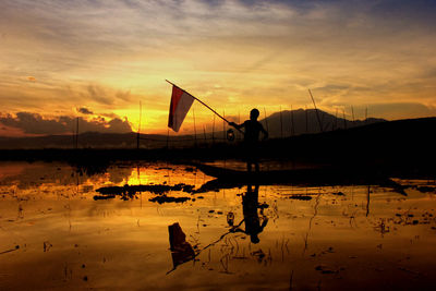 Silhouette man holding flag in lake against sky during sunset