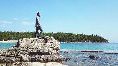 Man standing on rock at beach against clear blue sky