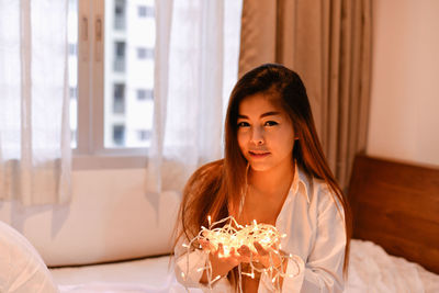 Portrait of smiling young woman with illuminated lights sitting on bed