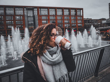 Young woman drinking water while standing by railing during winter