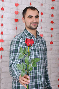 Portrait of smiling man proposing while holding red rose against decorated wall