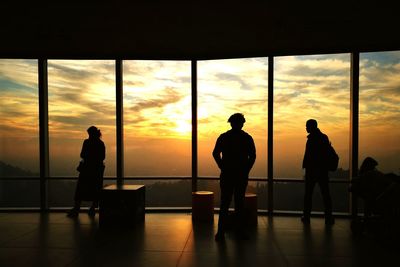 Silhouette people on floor against orange sky seen through window glass