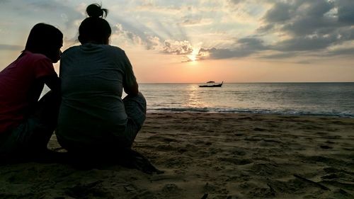 Rear view of women crouching on shore at beach against sky during sunset