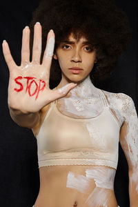 Portrait of woman with paint showing stop gesture with text while standing against black background