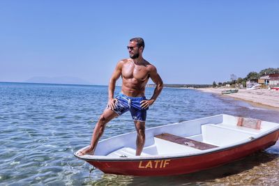 Full length of shirtless man standing on boat in sea against clear sky
