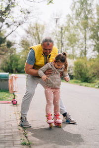 Full body of happy mature grandfather in casual clothes helping adorable positive girl practicing roller skating on asphalt asphalt road in forest