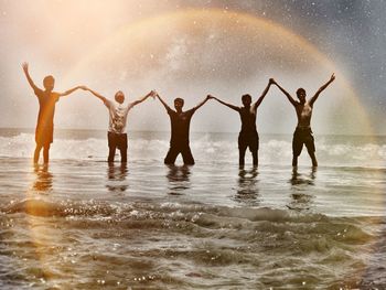 Male friends holding hands with arms raised while standing in sea against sky