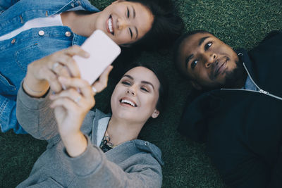 High angle view of smiling woman taking selfie with friends while lying on grass