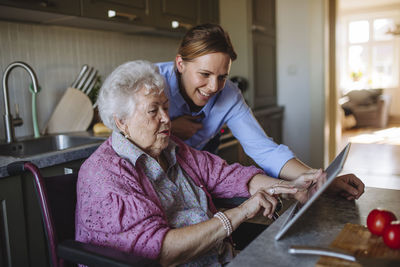 Happy home caregiver teaching tablet pc to senior woman at table