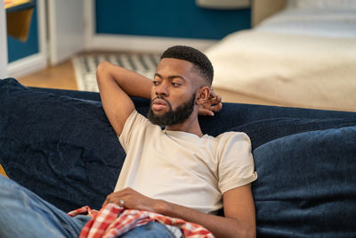 Thoughtful dreaming african american guy sits on sofa rests in studio apartment.