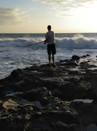 Rear view of man fishing while standing on rocks by sea during sunset