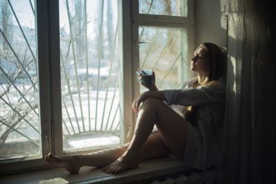 Full length of beautiful woman having coffee on window sill at home