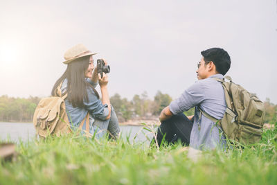 Young woman photographing man with camera while sitting on grassy field