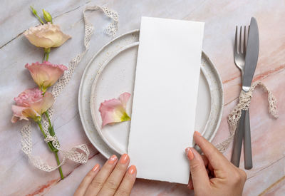 Hands holding wedding menu over a ceramic plate on a marble table decorated with flowers and ribbons