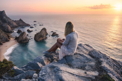 Woman sitting on rock by sea against sky during sunset