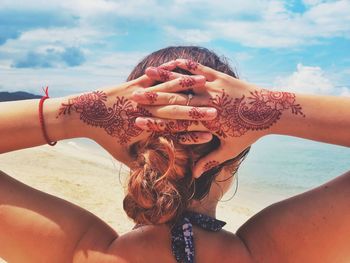 Rear view of young woman with hands behind head standing at beach against sky during sunny day