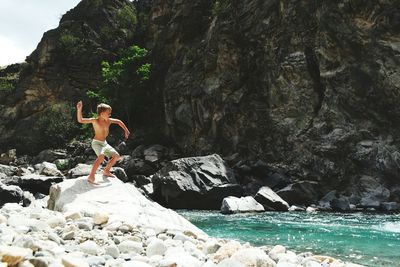 Full length of shirtless boy throwing stone in river while standing on rock